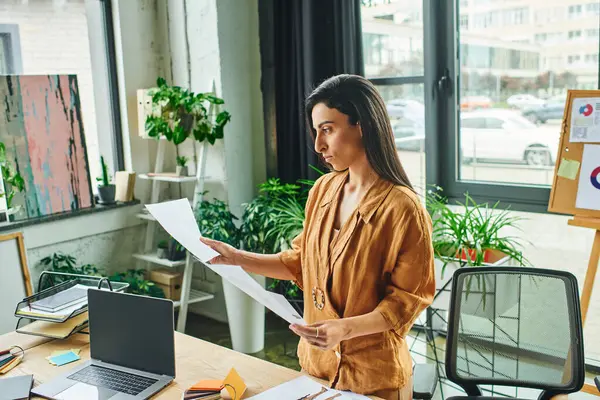 Una mujer en una oficina mira cuidadosamente un documento mientras está rodeada de plantas y un espacio de trabajo moderno. — Stock Photo