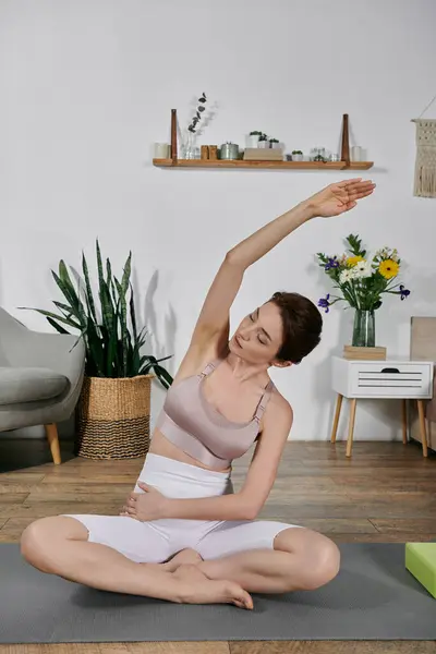 An Asian woman in a crop top sits cross-legged on a yoga mat, practicing a stretch with her arm extended overhead. — Stock Photo