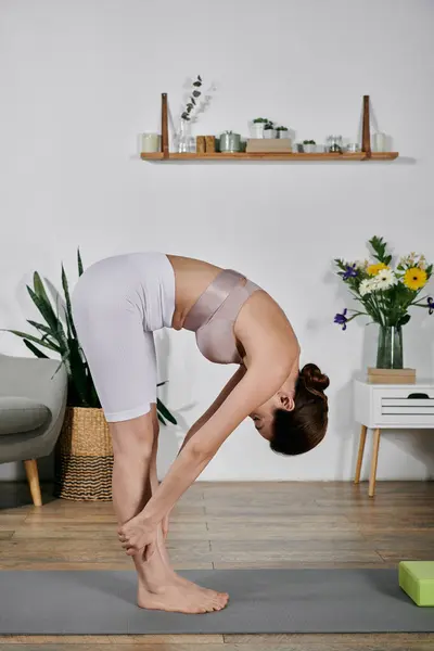 A young woman in a crop top bends forward, stretching her hamstrings on a yoga mat in her modern apartment. — Stock Photo