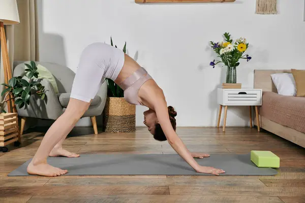 A woman in a white crop top and shorts performs a yoga pose on a gray mat in her living room. — Stock Photo