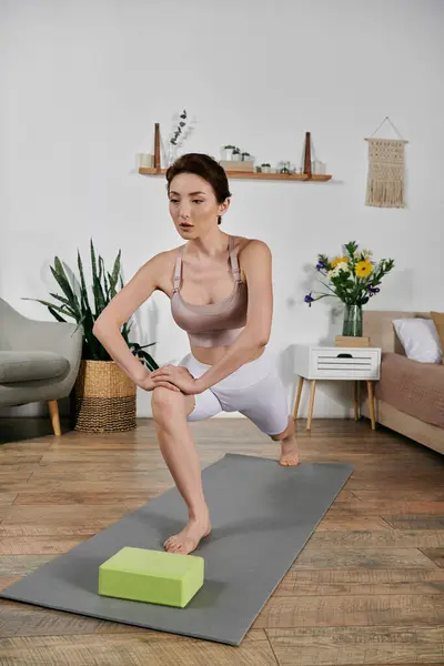 Eine Frau in einem bauchfreien Oberteil führt zu Hause eine Yoga-Stretchübung auf einer Matte durch, wobei sie einen Block zur Unterstützung benutzt. — Stockfoto