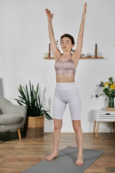 Young woman in crop top and leggings stands on a yoga mat in modern apartment with eyes closed, practicing meditation — Stock Photo