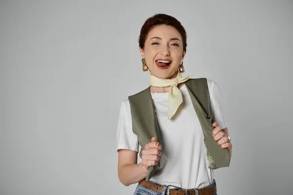 Una mujer asiática con estilo en un chaleco y cuello bufanda posa sobre un fondo gris, irradiando alegría y confianza. — Stock Photo