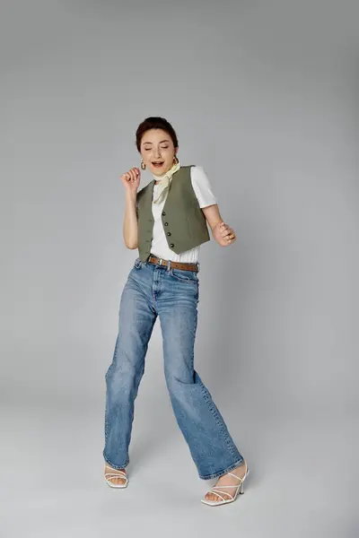 A stylish Asian woman in a green vest and white neck scarf poses against a gray background, wearing blue jeans and white sandals. — Stock Photo