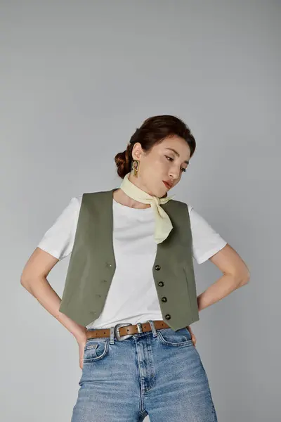 Une femme asiatique élégante dans un gilet vert et une écharpe jaune pâle pose sur un fond gris, ses cheveux tirés en arrière dans un chignon. — Photo de stock