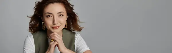 Una mujer asiática con el pelo castaño largo y un chaleco elegante posa sobre un telón de fondo gris, sus manos apretadas delante de ella. - foto de stock
