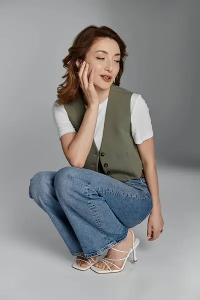 Stylish Asian woman in green vest and white t-shirt poses, hand on cheek, wearing blue jeans and white heels against a grey background — Stock Photo