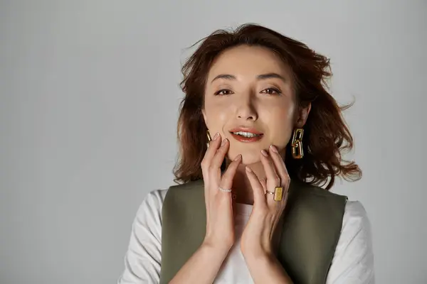 A stylish Asian woman in a vest poses against a gray backdrop, her hands delicately touching her face. — Stock Photo