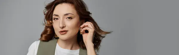 Una mujer asiática con estilo en un chaleco posa sobre un fondo gris, su mano suavemente tocando su pendiente. — Stock Photo