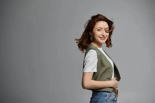 A beautiful woman with dark hair is dressed in a green vest and denim jeans. She smiles as she poses against a grey background. — Stock Photo