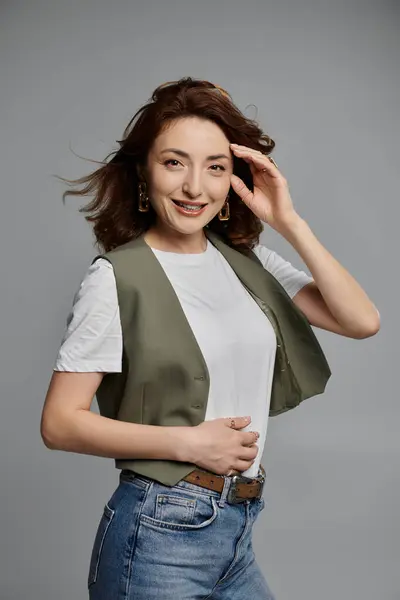 A stylish Asian woman with brown hair is wearing a green vest, a white t-shirt, and blue jeans, posing in front of a grey background. — Stock Photo