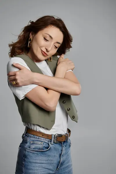 Une femme asiatique élégante portant un gilet pose sur un fond gris, les yeux fermés alors qu'elle s'embrasse. — Photo de stock