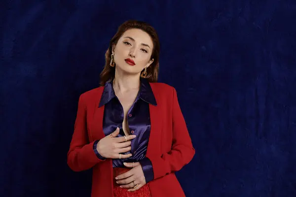 An attractive Asian woman poses in a red suit and silk shirt, her hands clasped in front of her, against a dark blue background. — Stock Photo