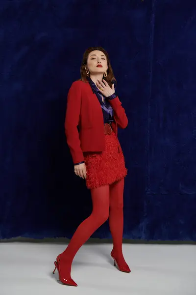 A woman in a red suit, silk shirt, and stockings poses against a blue wall. — Stock Photo
