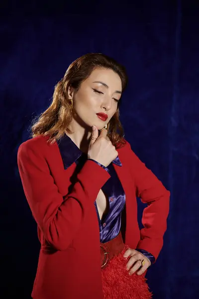 A stylish Asian woman in a red suit and silk shirt poses with one hand on her chin against a dark blue background. — Stock Photo