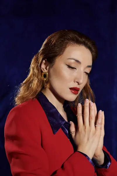 A stylish Asian woman in a red suit and silk shirt poses with her hands clasped in front of her, against a dark blue background. — Stock Photo
