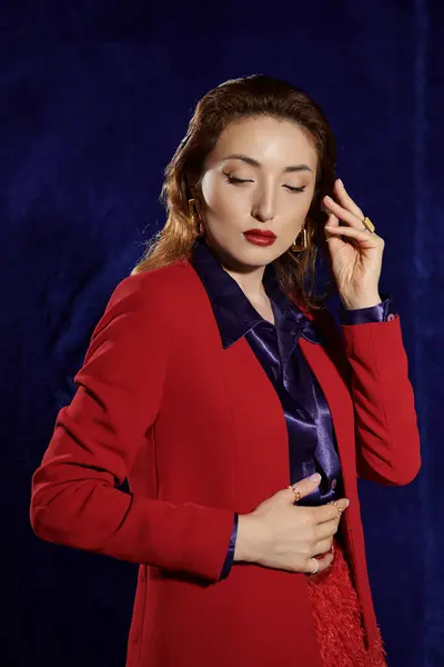 A stylish Asian woman poses in a crimson suit and silk shirt, her gaze lost in thought, against a dark backdrop. — Stock Photo