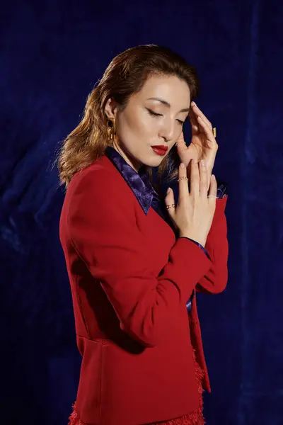 A woman with long brown hair wears a red suit and silk shirt, posing with her hands clasped in front of her against a deep blue background. — Stock Photo