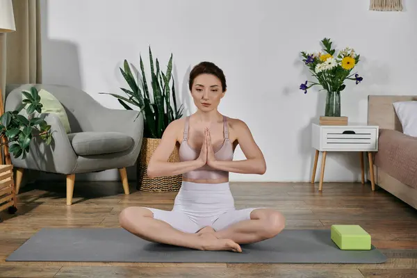 Una mujer se sienta en una pose de yoga sobre una esterilla, con las manos en una posición de oración, meditando en un apartamento moderno. - foto de stock