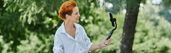 A redhead films video in park with selfie stick, smiling — Stock Photo