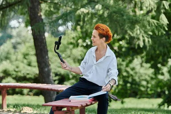 A woman in a white shirt sits on a bench outside and uses a selfie stick to record herself. — Stock Photo