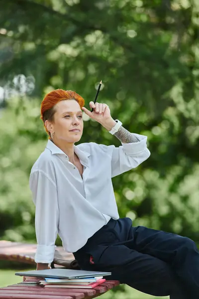Red-haired teacher in white shirt, black pants, sitting on bench lost in thought with pen — Stock Photo