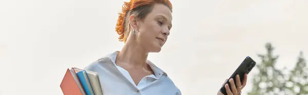 A teacher with red hair checks her phone, holding textbooks. — Stock Photo