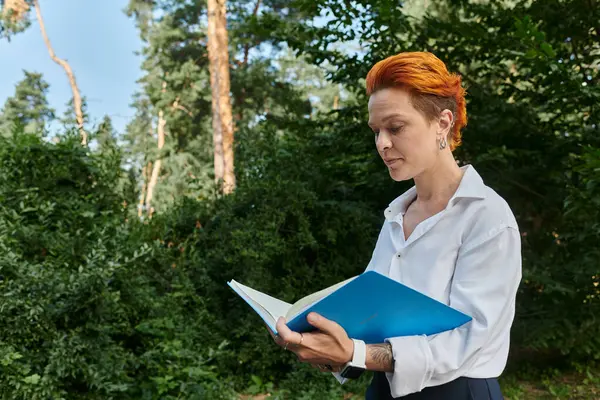 A woman with fiery red hair reads a book in a wooded area on campus. — Stock Photo