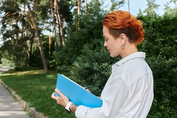 Жінка з рудим волоссям стоїть на кампусній доріжці, роблячи нотатки в синьому зв'язувачі. — стокове фото