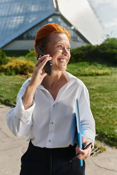 A woman with bright red hair smiles while speaking on her phone, holding a folder. — Stock Photo