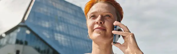 Woman with red hair smiles, talks on phone with campus building in background — Stock Photo