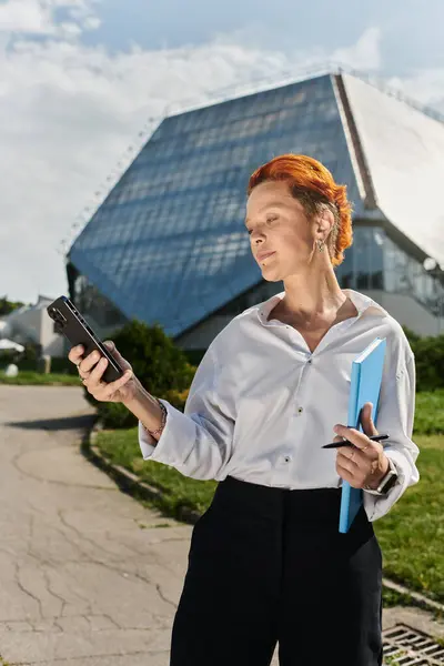 Une femme aux cheveux courts roux traverse le campus, tenant un téléphone et un dossier bleu. — Photo de stock