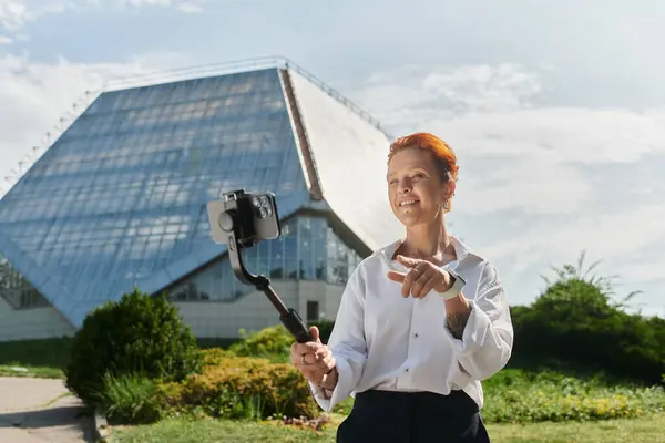 A woman records a video on her phone while standing in front of a modern campus building. — Stock Photo