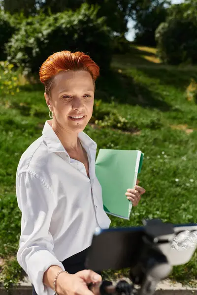 A smiling teacher with red hair stands in the park, holding a folder and a camera. — Stock Photo