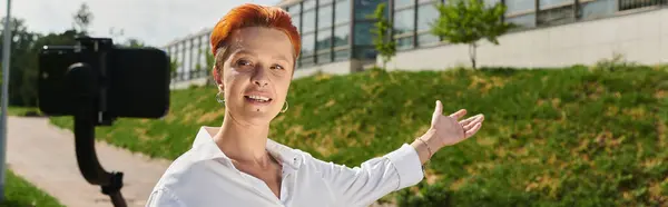 A teacher with red hair smiles and gestures while filming a vlog outside a campus building. — Stock Photo