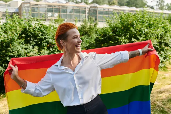 A woman smiles while holding a rainbow pride flag on a campus. — Stock Photo