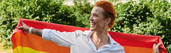 A woman with red hair and a big smile holds up a pride flag on a sunny campus. — Stock Photo