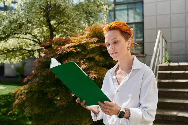 A woman with bright orange hair reads a book on a sunny campus. — Stock Photo