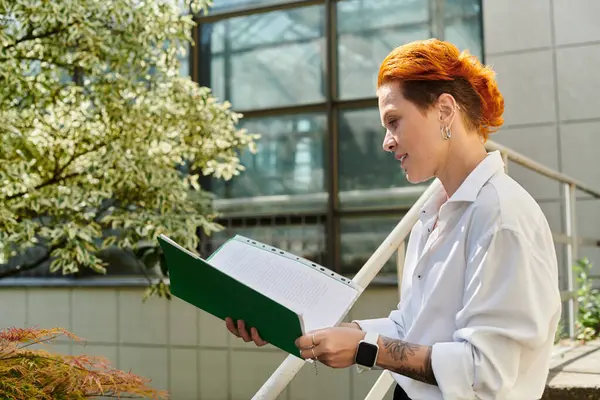 A woman with vibrant red hair reads a document outside on campus. — Stock Photo