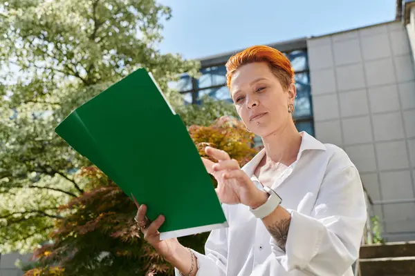 Una donna con i capelli rossi corti recensioni note su una cartella verde mentre in piedi all'aperto nel campus. — Foto stock