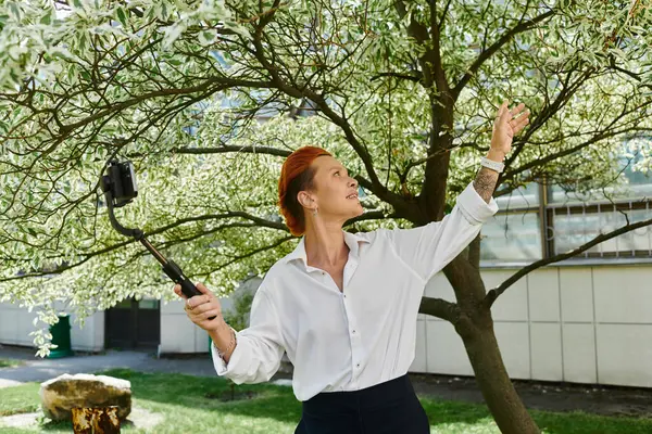 A woman with red hair takes a selfie with a selfie stick in front of a tree. — Stock Photo