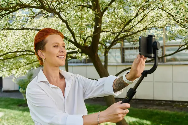A woman in a white shirt smiles while taking a selfie with a phone on a tripod. — Stock Photo