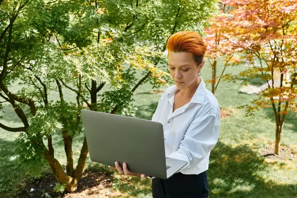 A teacher with short, fiery hair works on her laptop, enjoying the peace of a campus courtyard. — Stock Photo