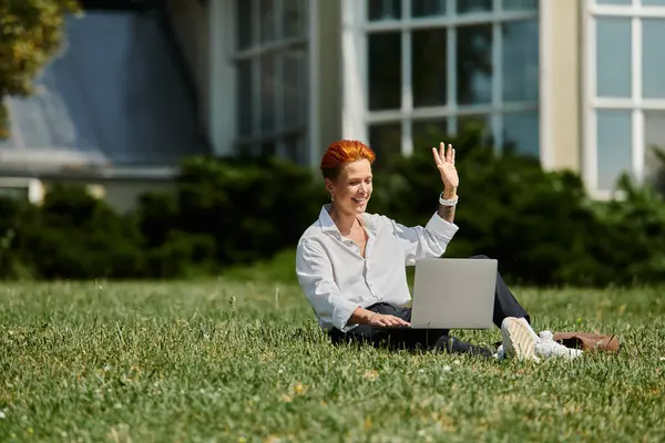 A teacher sits on the grass outside on a sunny day, waving and working on a laptop. — Stock Photo