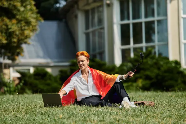 A woman wearing a rainbow flag sits on a grassy campus lawn, using a laptop. — Stock Photo