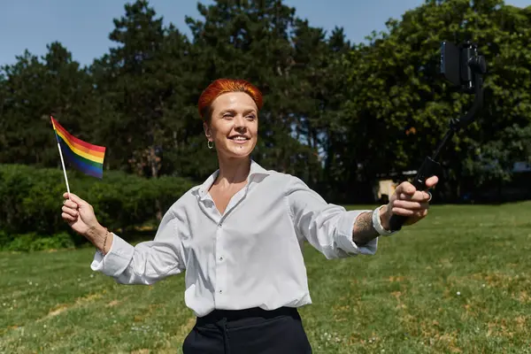 Woman with rainbow flag takes selfie on campus with phone on selfie stick — Stock Photo