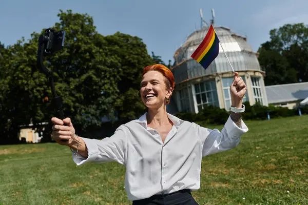 A smiling woman holds a rainbow flag and takes a selfie outside a campus building. — Stock Photo