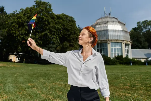 A woman with red hair holds a rainbow flag on a sunny day in a campus setting. — Stock Photo
