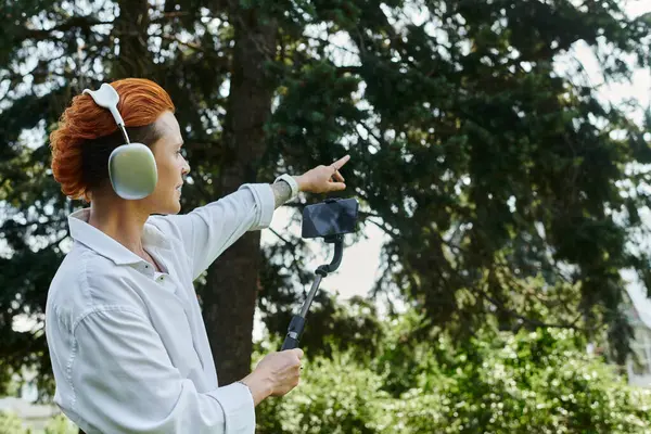 A woman wearing headphones films with a selfie stick in a campus setting. — Stock Photo