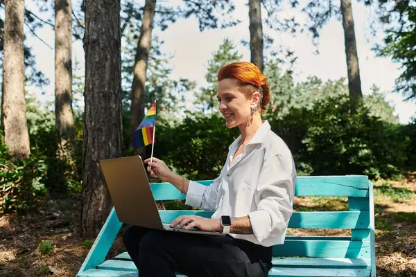 A woman sits on a bench in a wooded campus, holding a rainbow flag and using a laptop. — Stock Photo